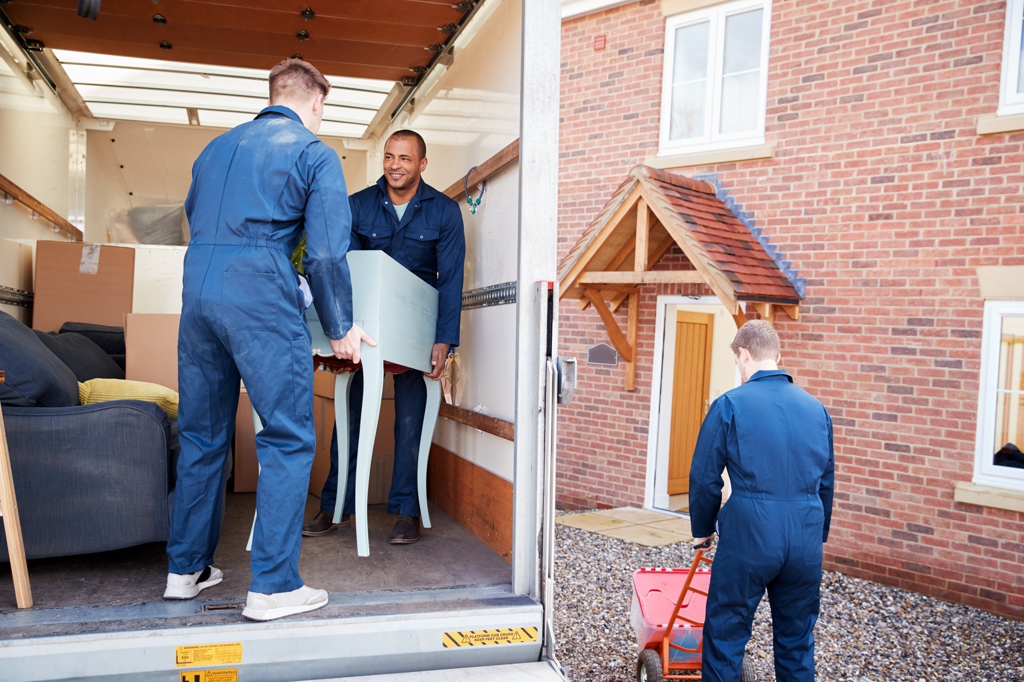 Removal Company Workers Unloading Furniture And Boxes From Truck Into New Home On Moving Day